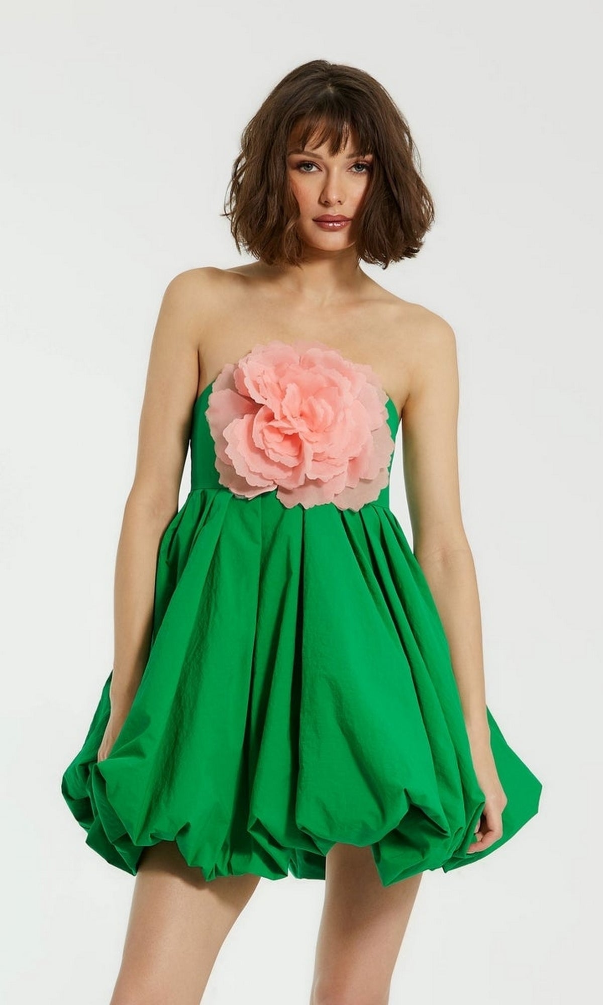 A woman poses against a plain background wearing the Mac Duggal 20931 short bubble party dress—strapless, green with a balloon hem and a bold pink flower accent, ideal for any semi-formal event.